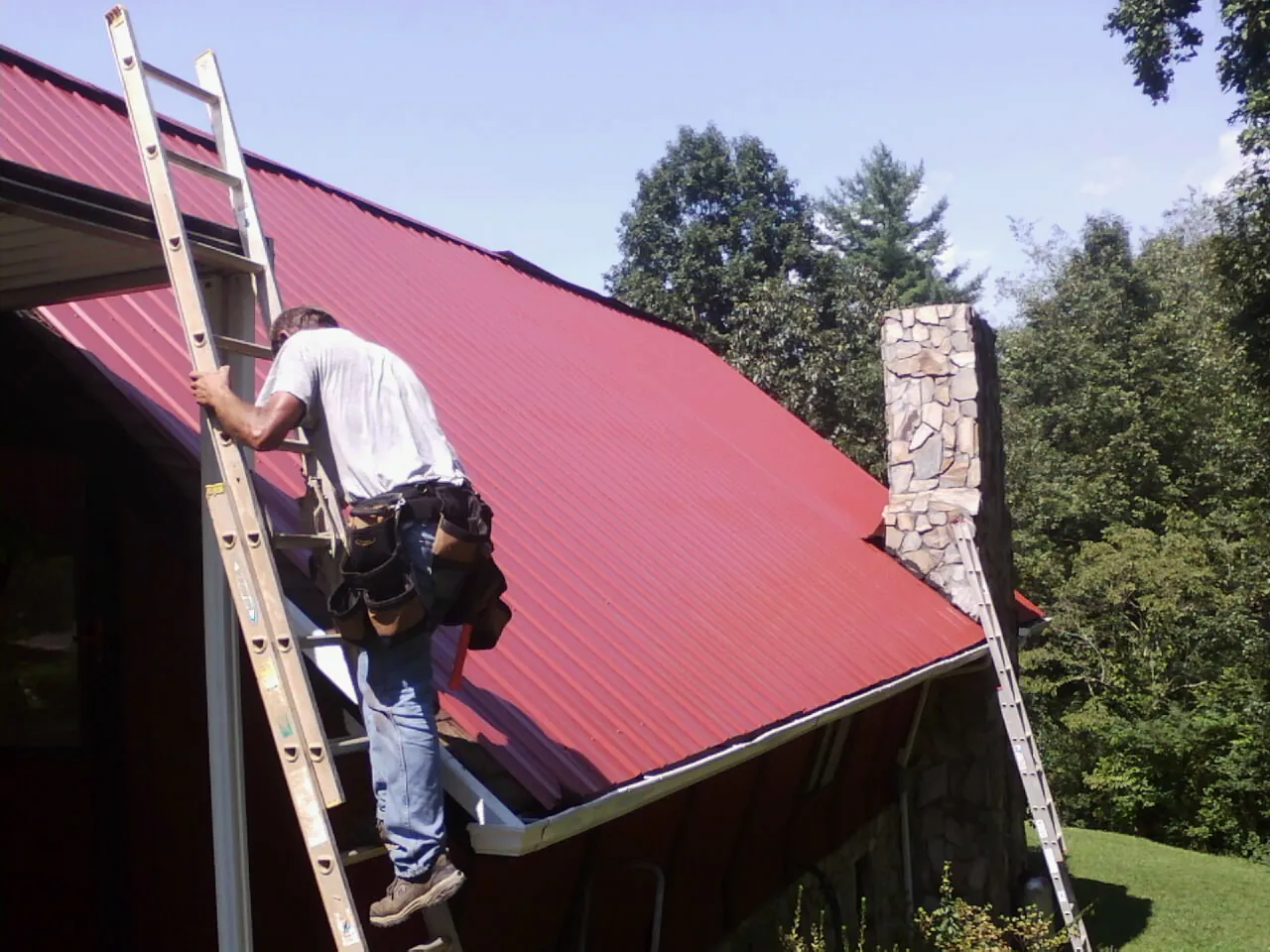 Roofer on a ladder beside a red metal roof and stone chimney — Soffit & Fascia Repair by Roofing Brothers in Taft